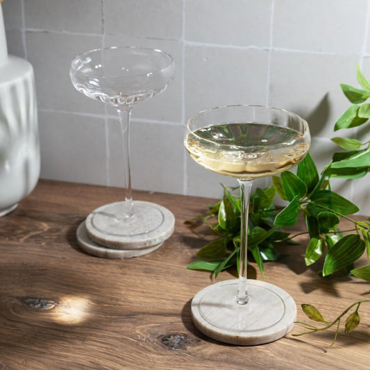 Two coupe glasses with champagne on a wooden surface with a plant in the background