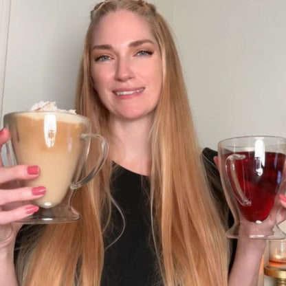 Woman holding two mugs, one with a creamy beverage and the other with a red liquid, against a neutral background.