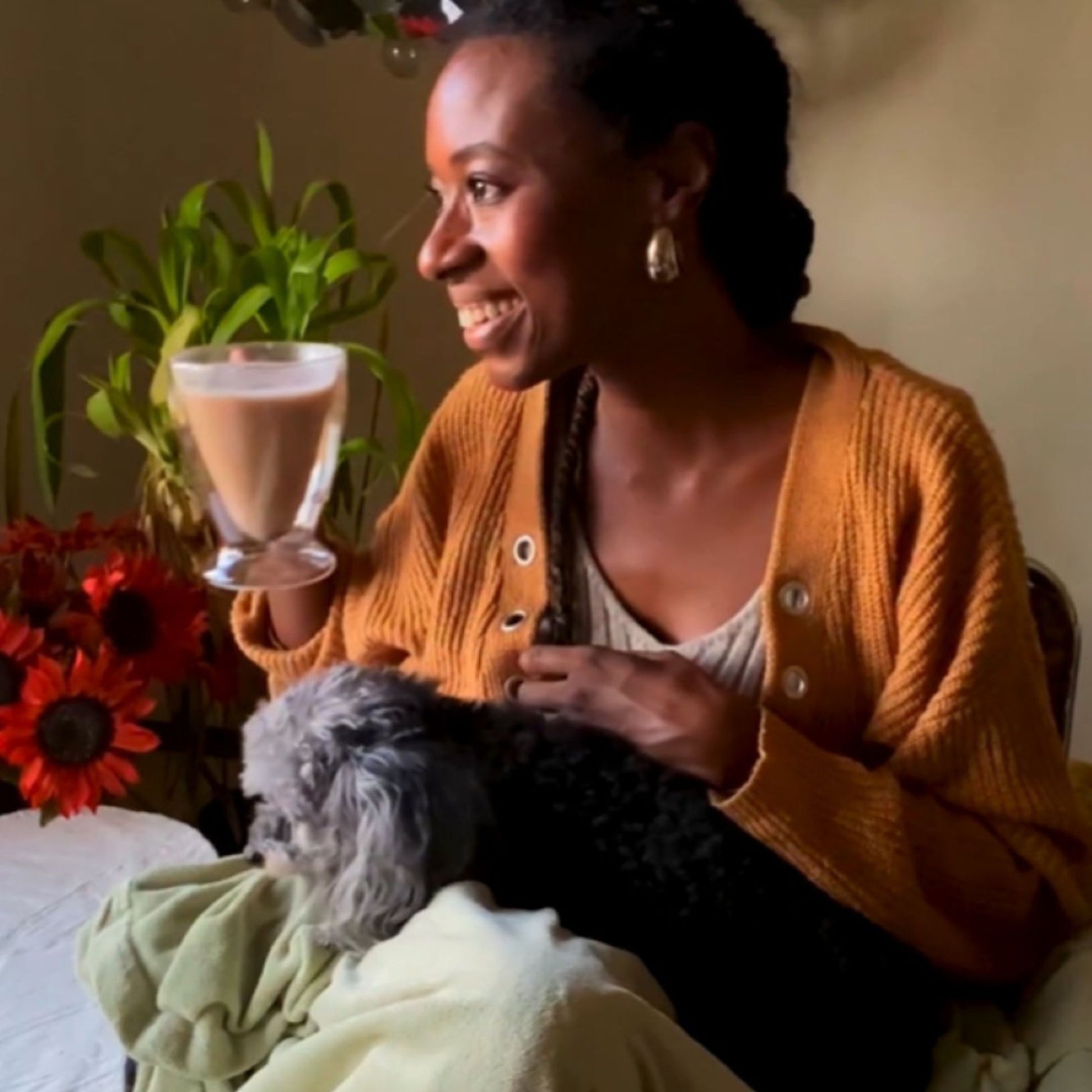 Woman holding a glass of chocolate milk and a small dog, surrounded by plants and flowers.