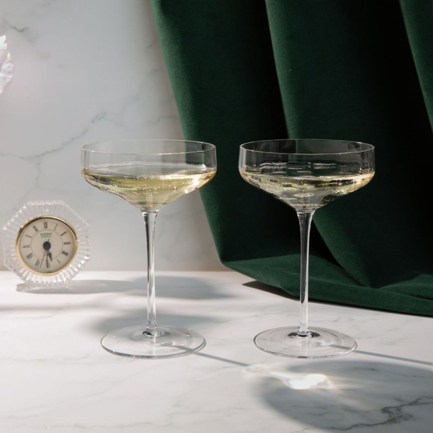 Two champagne glasses with a small clock on a marble surface with green fabric in the background.