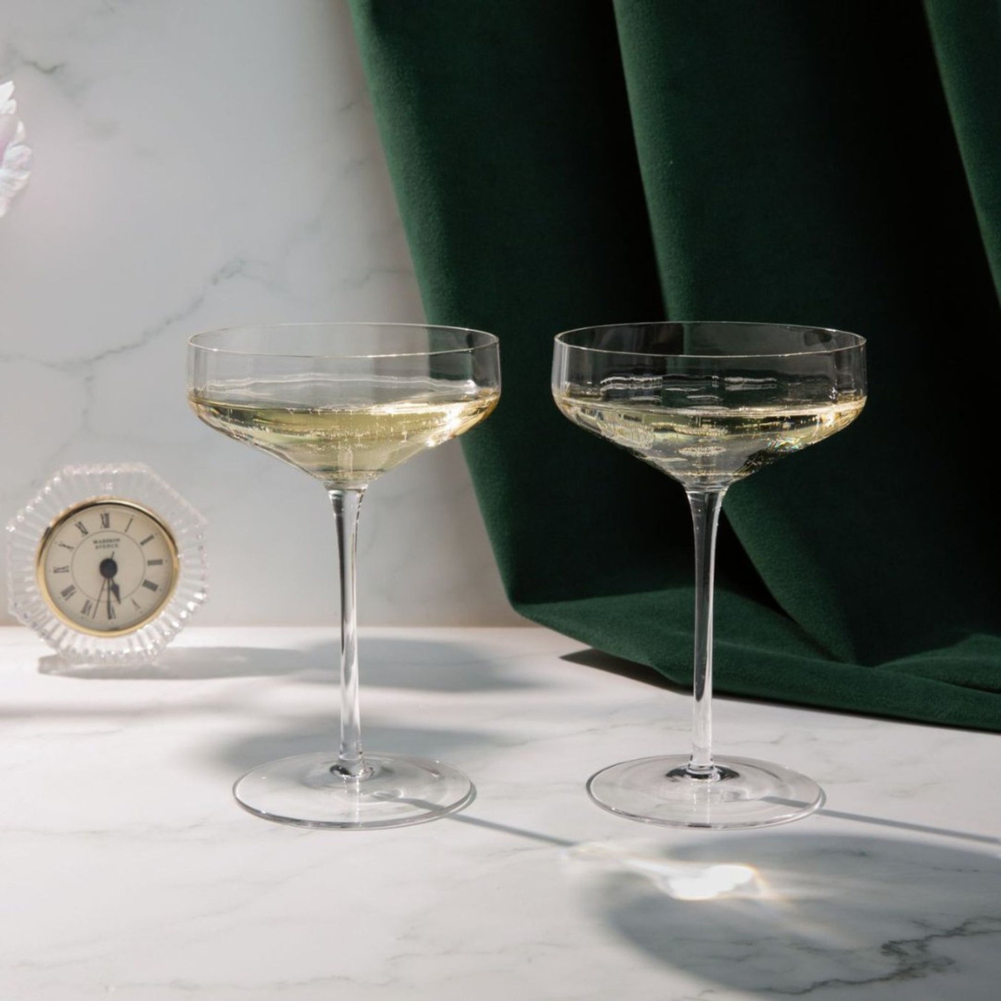 Two champagne glasses with a small clock on a marble surface with green fabric in the background.