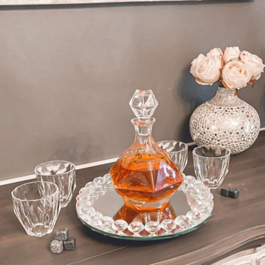 Glass decanter with amber liquid on a mirrored tray, surrounded by glasses and a decorative vase with flowers.