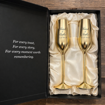 Two gold champagne flutes in a black box with white interior, engraved with 'Trish & Gabe' and a date.