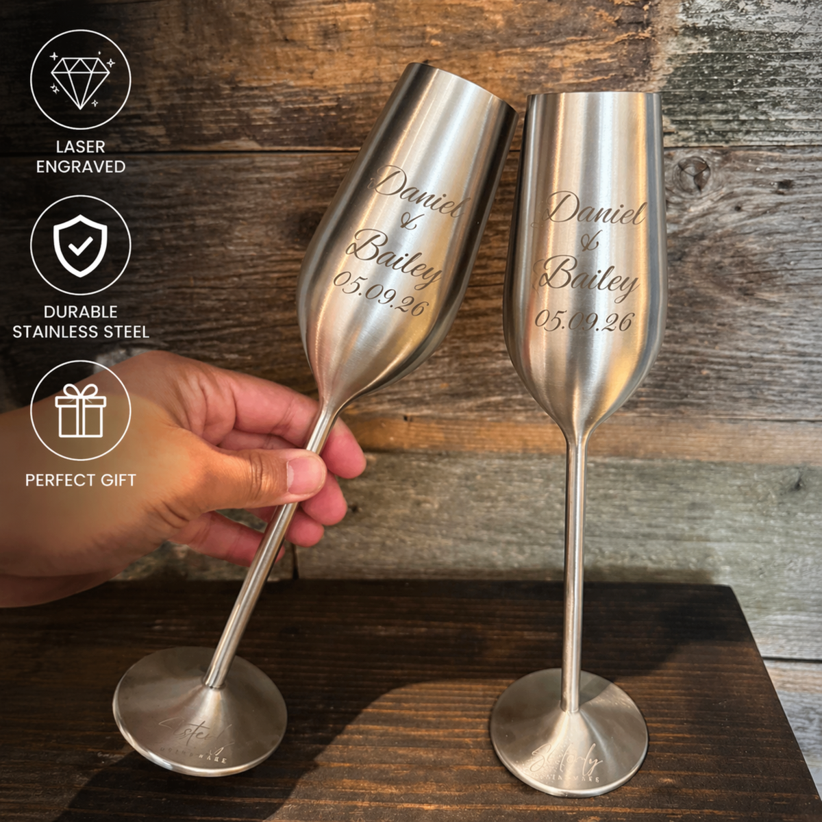 Two silver champagne flutes with engraved text on a wooden surface, held by a hand.