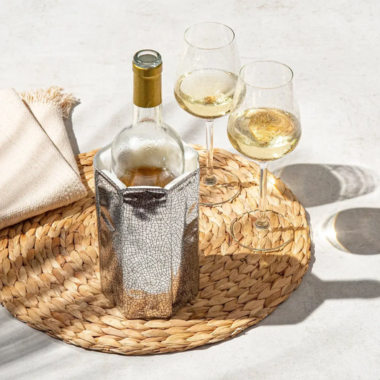 Bottle of white wine in a decorative holder with two glasses on a woven mat.