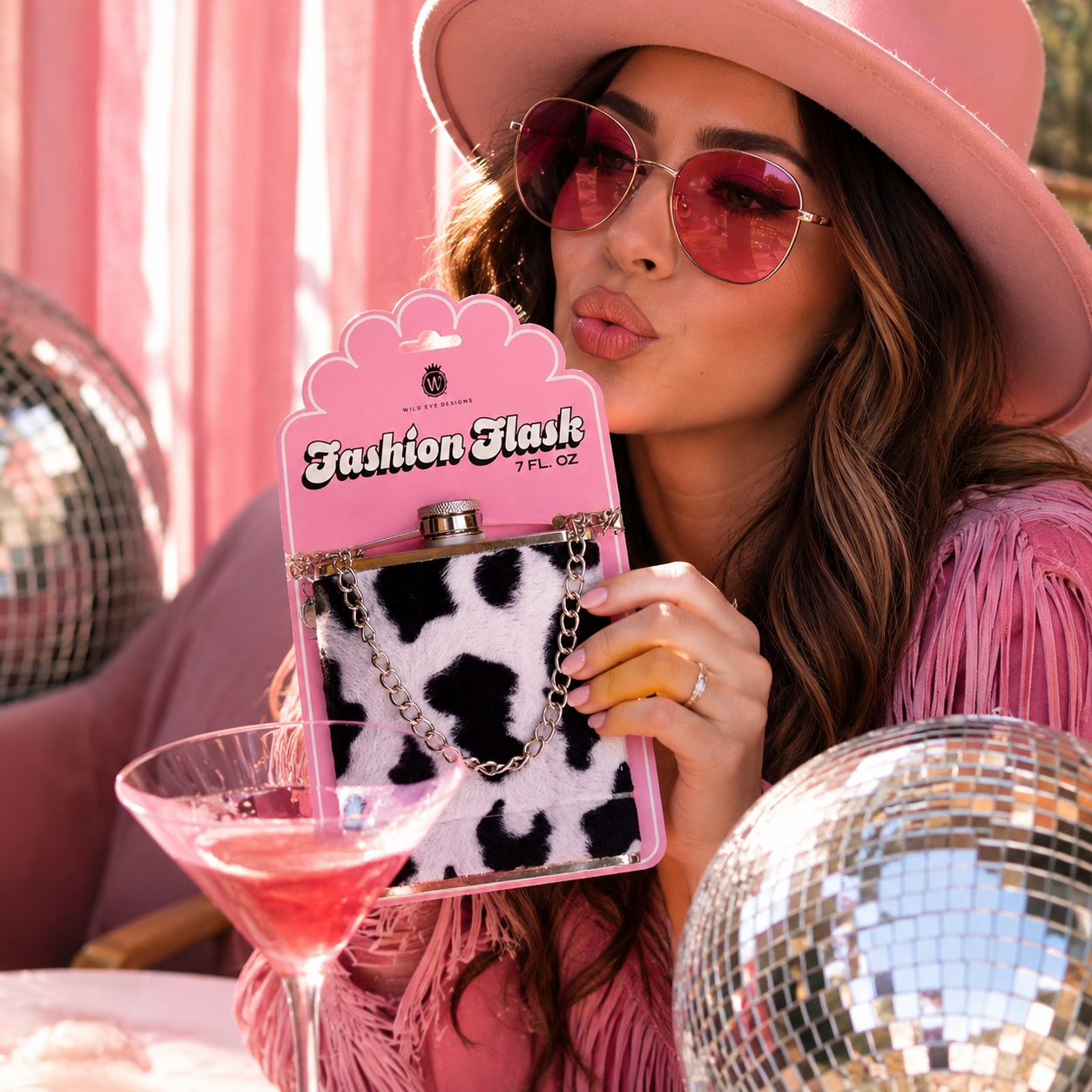 A woman in a pink hat and sunglasses holds a pink package containing a small black-and-white cow-print flask with a chain strap. Disco balls and a pink drink are visible in a party-like setting.
